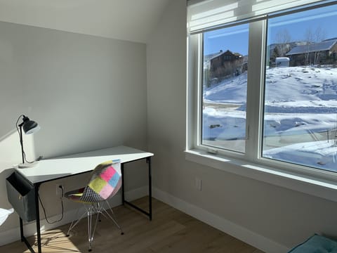 A neat, minimalistic workspace with a desk, lamp, and colorful chair next to a large window with a snowy landscape outside.