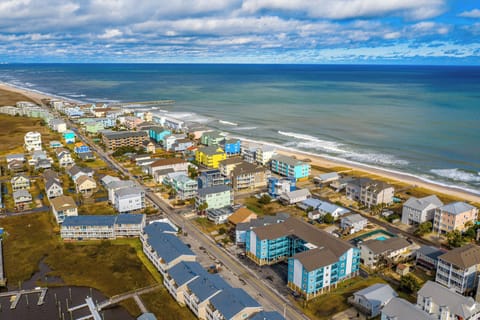 Colorful coastal community stretches along pristine beach with rolling waves and expansive ocean views under dramatic cloudy skies.