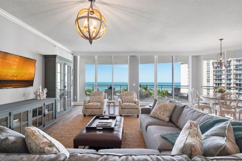The living room features gray sofas, floral armchairs, a leather ottoman, and floor-to-ceiling windows overlooking the ocean.