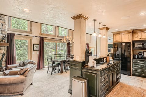 Open-concept living and dining area with large windows, stone fireplace, and high ceilings. Kitchen island with black countertop and pendant lights. Dark green cabinets and stainless steel appliances.