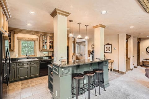 A kitchen with green cabinets, granite countertops, three bar stools, and stainless steel appliances. There is recessed lighting and a window above the sink.
