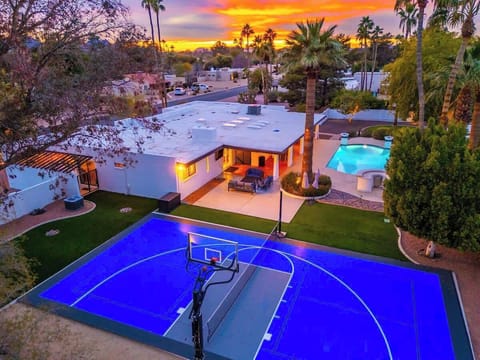 Aerial view of a desert vacation property featuring a private basketball court, heated pool, and expansive outdoor entertaining spaces, set among palm trees with spectacular sunset skies overhead.