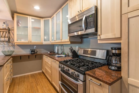 A modern kitchen with stainless steel appliances, including a microwave and gas stove, light wood cabinetry, glass-front upper cabinets, and a brown speckled countertop.