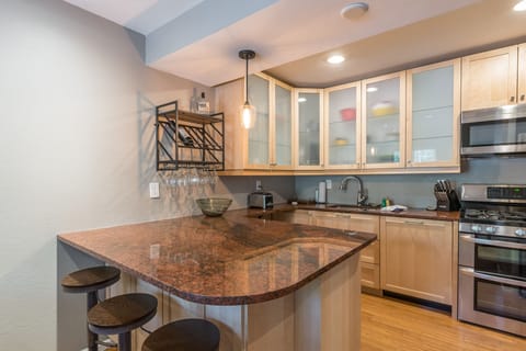A modern kitchen features light wood cabinetry, stainless steel appliances, a granite countertop with a breakfast bar and three stools, and a hanging wine rack with glasses.