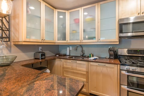 A modern kitchen featuring light wood cabinets with frosted glass doors, granite countertops, stainless steel appliances, and a sink with a pull-down faucet. A toaster and knife block are also visible.
