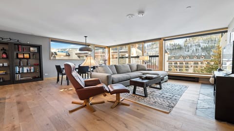 Relax in your spacious living room with floor-to-ceiling windows framing stunning mountain views. Natural light fills the open space, highlighting plush seating and warm wood floors that invite you to unwind after a day of adventure.