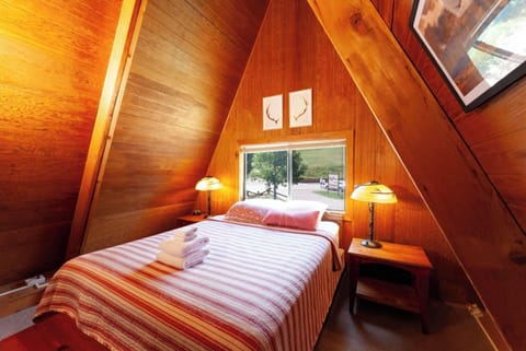 Cozy A-frame bedroom with rich wood paneling and warm lighting. Your plush bed sits beneath a charming window, offering peaceful views of the surrounding landscape.
