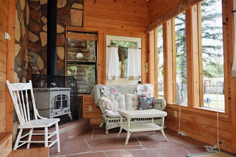 Curl up in the cozy wicker loveseat by the wood-burning stove, where floor-to-ceiling windows frame peaceful forest views and warm pine walls wrap you in rustic charm.