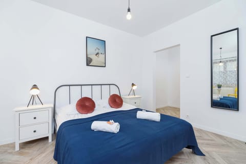 A spacious and elegant bedroom featuring a large double bed dressed in navy blue bedding. The room is bright, with white bedside tables, lamps, and a decorative piece on the wall adding to its charm.