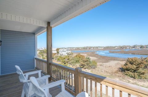 Covered balcony with white chairs, wood railing, blue siding, clear sky, marsh grass, waterway, trees, and scenic Oak Island, NC coastal views.