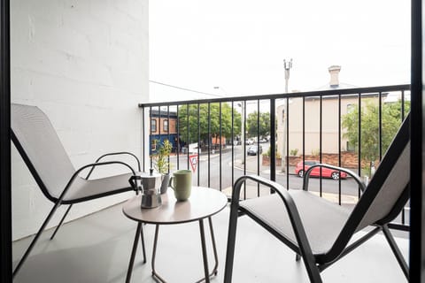 A cozy apartment balcony featuring two chairs, a small table with coffee, and a view overlooking a city street.
