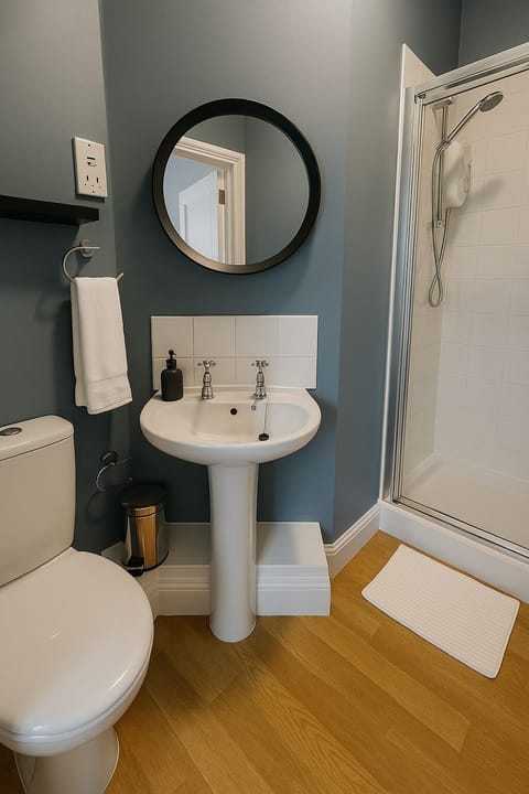Cosy bathroom with elegant blue-gray walls and a sleek white pedestal sink. Features a modern electric shower, a minimalist round mirror, and shaving socket, ideal for a relaxing break. 