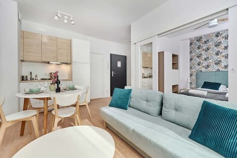 A bright and modern living room with a light wooden floor. It features a comfortable teal-colored sofa, a white coffee table, and a wall-mounted television. Decorative elements and natural light add to its coziness.
