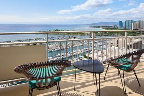Expansive balcony showcasing a stunning marina dotted with sailboats, sandy beaches stretching along turquoise waters, and city skyline framed by mountains. Perfectly positioned for taking in Hawaii's coastal beauty.
