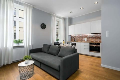 A bright living room with a gray sofa, a small round table, and a view into the kitchen area.