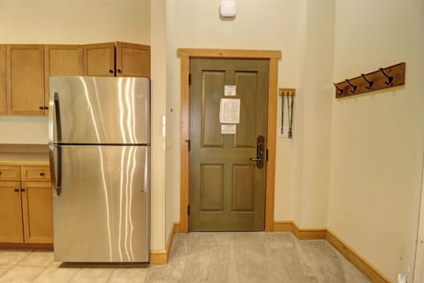 A kitchen area with wooden cabinets and a stainless steel refrigerator next to a green door. The door has papers taped to it and a wall-mounted coat rack with hooks is on the right.