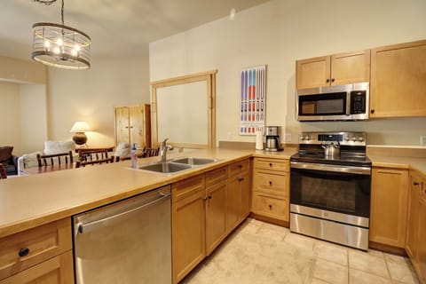 A modern kitchen with wooden cabinets, stainless steel appliances including an oven and dishwasher, and a double sink. A colorful wall art piece is visible above the countertop.