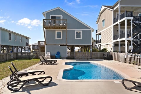Oak Island, NC rental with private pool, lounge chairs, fenced yard, elevated blue beach house, balconies, grill, patio, and sunny coastal setting.