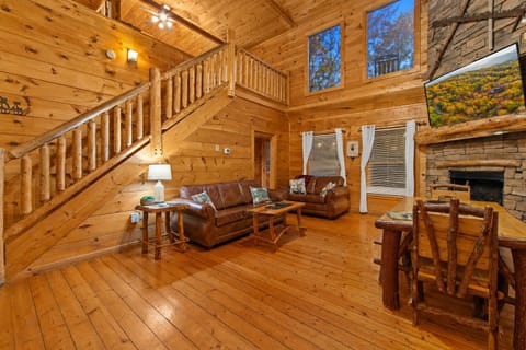 This open-concept cabin living area boasts cozy leather couches, a rustic wooden staircase, and a fully equipped kitchen with black appliances, all set within a warm, wood-paneled interior.