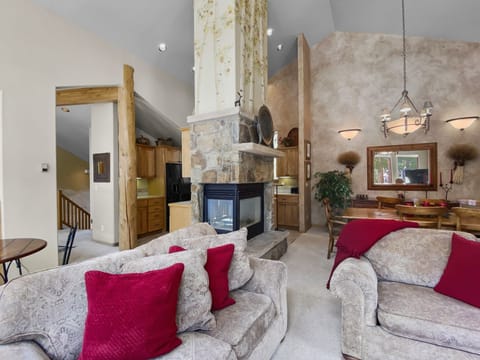 A living room with beige sofas and red cushions, a stone fireplace, and an adjacent dining area featuring a wooden table and chandelier. The room has high ceilings and neutral-toned walls.