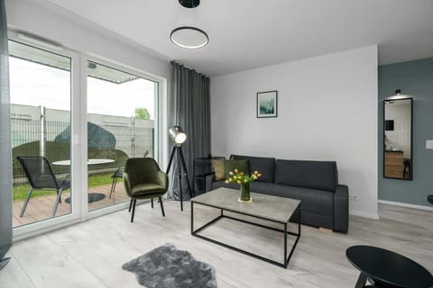 A modern living room with minimalistic design featuring a comfortable grey sofa, a glass coffee table, a floor lamp, and large windows allowing natural light to flood the space, with a view of a green garden.