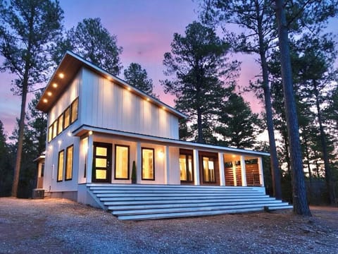A modern barn-style retreat glows warmly at dusk, nestled among tall pines with a painted sky behind. The wide front steps and wrap-around deck invite a peaceful arrival in nature.