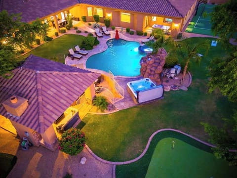 An aerial view showcasing the property's impressive outdoor amenities at twilight. The illuminated pool, hot tub, landscaped gardens, and miniature golf course create a resort-like private backyard setting.