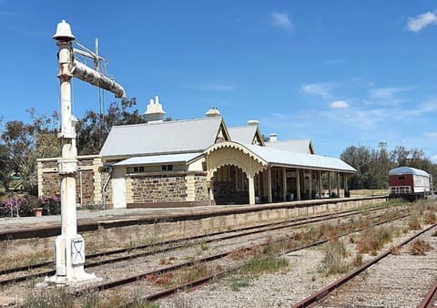 A historic stone railway station with ornate roofing and a wide platform stands beside old, overgrown train tracks under a bright blue sky, with a single red and white train car in the distance.