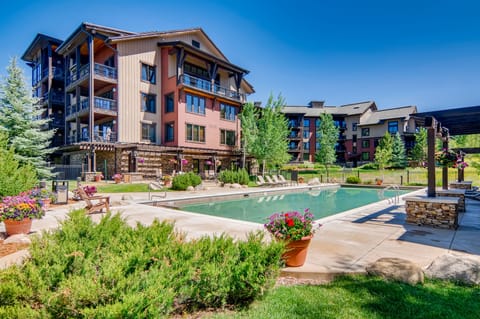 Outdoor swimming pool with surrounding lounge chairs, manicured greenery, and potted plants. Multi-story residential buildings in the background under a clear blue sky.