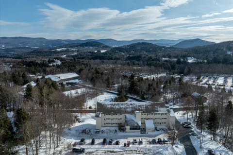 Aerial winter view of the Village Gate Building nestled in a snow-covered mountain valley with forested hills and scenic landscape stretching into the distance.