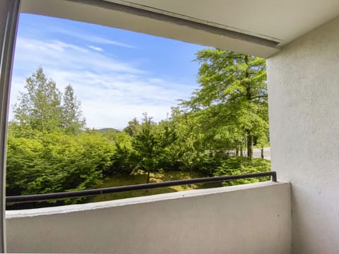 A daylight view from a covered balcony overlooks trees and shrubbery with a road visible in the background. The sky is clear with some clouds.