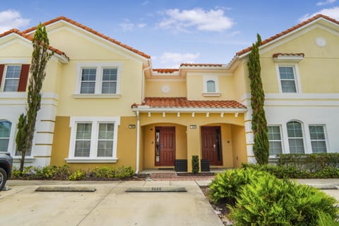 Yellow stucco townhome with red tile roof, twin front doors, and trimmed landscaping in parking area.