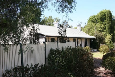 This image shows a charming, rustic cottage surrounded by lush greenery and partially enclosed by a white picket fence. The building features a long, sloped roof with chimneys and a shaded veranda, evoking a peaceful, countryside ambiance.