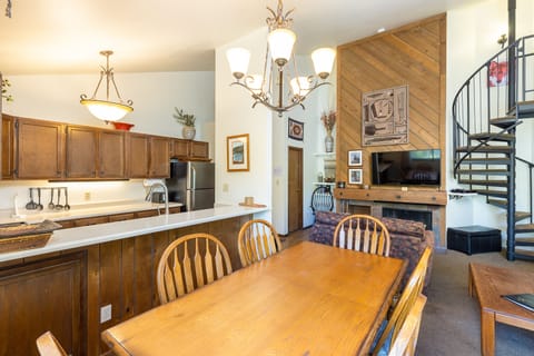 A dining area with a wooden table and chairs connects to a kitchen with wooden cabinets and stainless steel appliances. A nearby living space has a fireplace, a TV, and a spiral staircase.