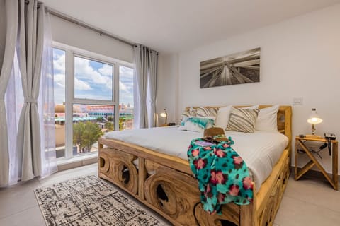 Wake up to natural light streaming through floor-to-ceiling windows offering colorful rooftop views. Your cozy retreat features a handcrafted wooden bed dressed in crisp linens, bedside lamps for evening reading, and bright, welcoming decor.
