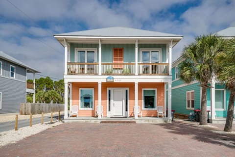Charming two-story beach house with coral and aqua exterior, welcoming front porch, and tropical palm trees in a coastal neighborhood.