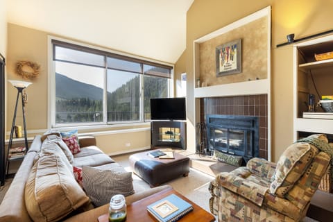 A cozy living room with a large window showcasing a mountain view, a TV beside a fireplace, a beige sofa, patterned armchair, and a glass jar on a small table. Bookshelves are built into the wall.
