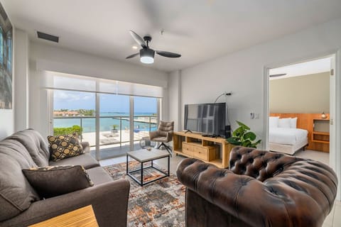 Unwind in your stylish living area with stunning turquoise water views through floor-to-ceiling windows. A coastal breeze flows through as you relax in plush seating.