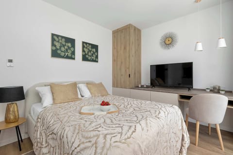 A cozy studio-style bedroom features a double bed with patterned bedding, a small desk area, and a compact kitchenette along one wall. Soft beige tones and wood finishes create a warm atmosphere.