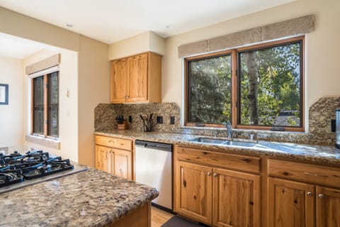 A kitchen with granite countertops, wooden cabinets, a gas stove on an island, a sink under a window, and a dishwasher.