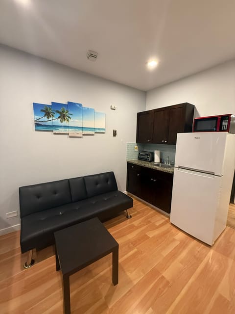 This inviting living area doubles as a compact kitchenette, perfect for relaxing or enjoying a light meal at home. The space features a sleek black futon sofa, ideal for lounging or accommodating an extra guest, complemented by a vibrant beach-themed