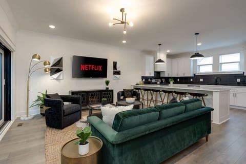 Living room with emerald green velvet sofa, Netflix display, kitchen island, and pendant lights