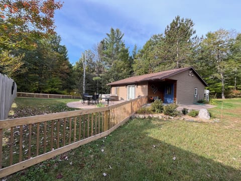 A small, single-story brown cabin with a white door and a large concrete patio, surrounded by a green lawn and tall pine trees.