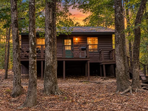 A cozy log cabin nestled among towering trees, with warm lights glowing through the windows as autumn leaves blanket the forest floor and a vibrant sunset paints the sky.