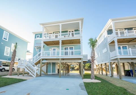 Tall coastal home with blue siding, white balconies, covered parking, exterior stairs, palm trees, concrete driveway, and bright beach neighborhood views.