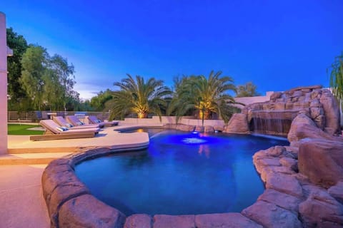 A stunning resort-style pool features dramatic rock formations, a cascading waterfall, and illuminated palms—perfect for evening relaxation under the open sky.