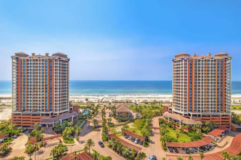 Aerial view showing twin resort towers framing the pristine white sand beach and emerald Gulf waters beyond, with lush landscaping and parking facilities completing the property's coastal entrance.