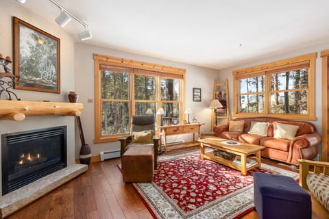 A cozy living room with a leather sofa, armchair, a wooden coffee table, a fireplace on the left, large windows with wooden frames, and a red patterned rug on a wooden floor.