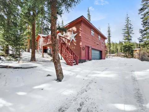 A red wooden cabin with a garage stands in a snowy landscape, surrounded by pine trees. A staircase leads to the front door, and a large white star decorates the side of the house.