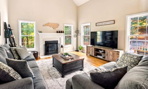 Gather in the light-filled living room with vaulted ceilings, cozy seating, and a central fireplace overlooking peaceful wooded views.